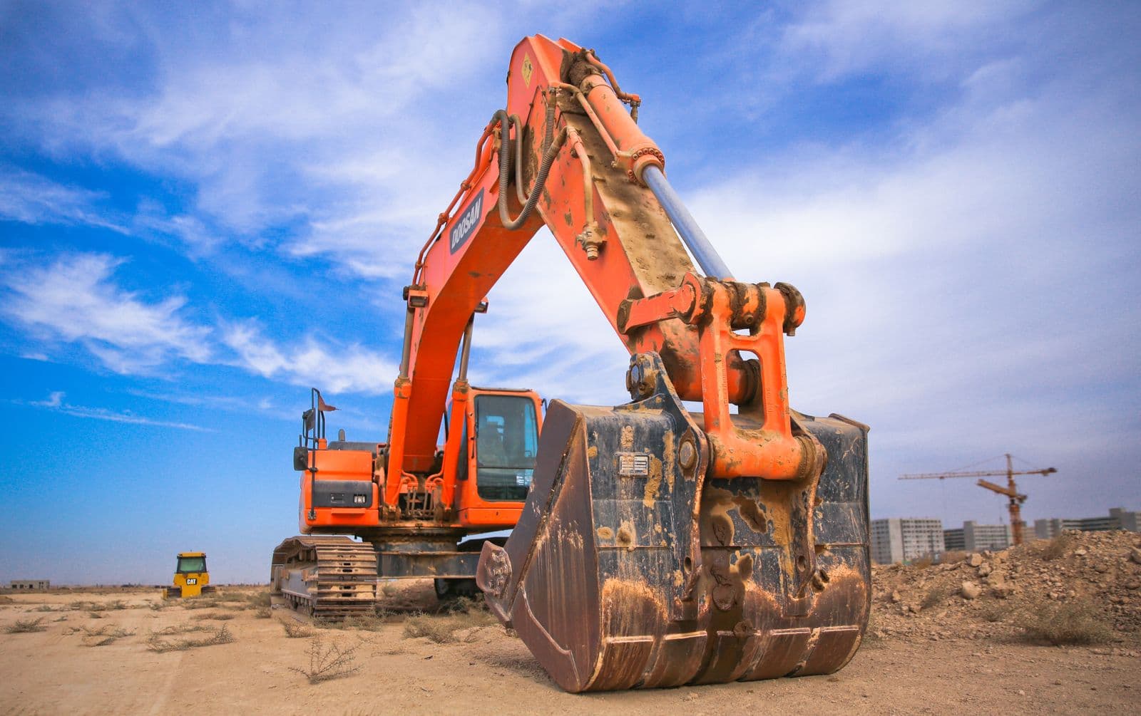 Orange excavator on a dusty construction site with cranes in the distance